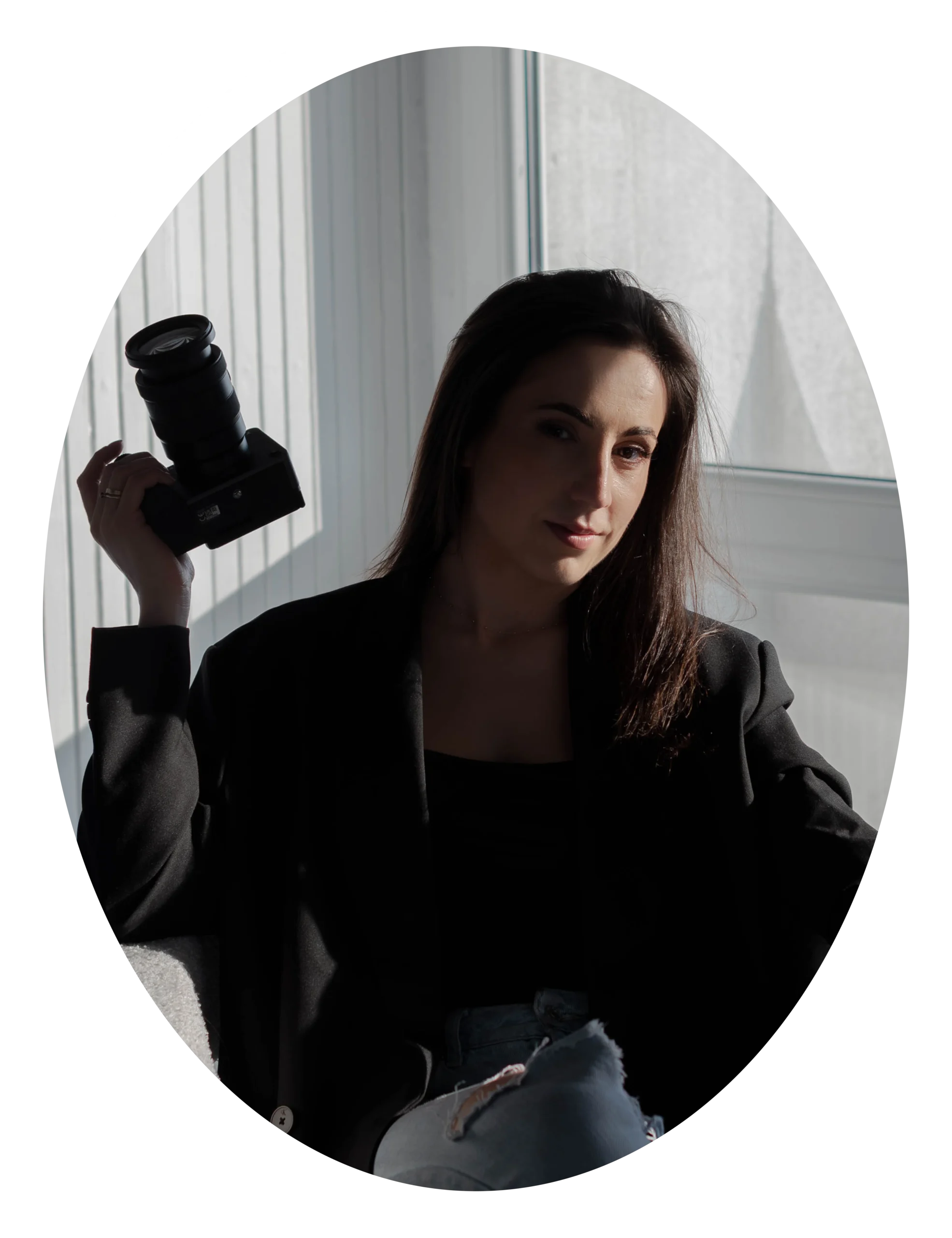 Młoda kobieta w ciemnych włosach siedzi na fotelu w półcieniu. Ma na sobie czarną marynarkę i jeansy. W dłoni trzyma aparat fotograficzny.