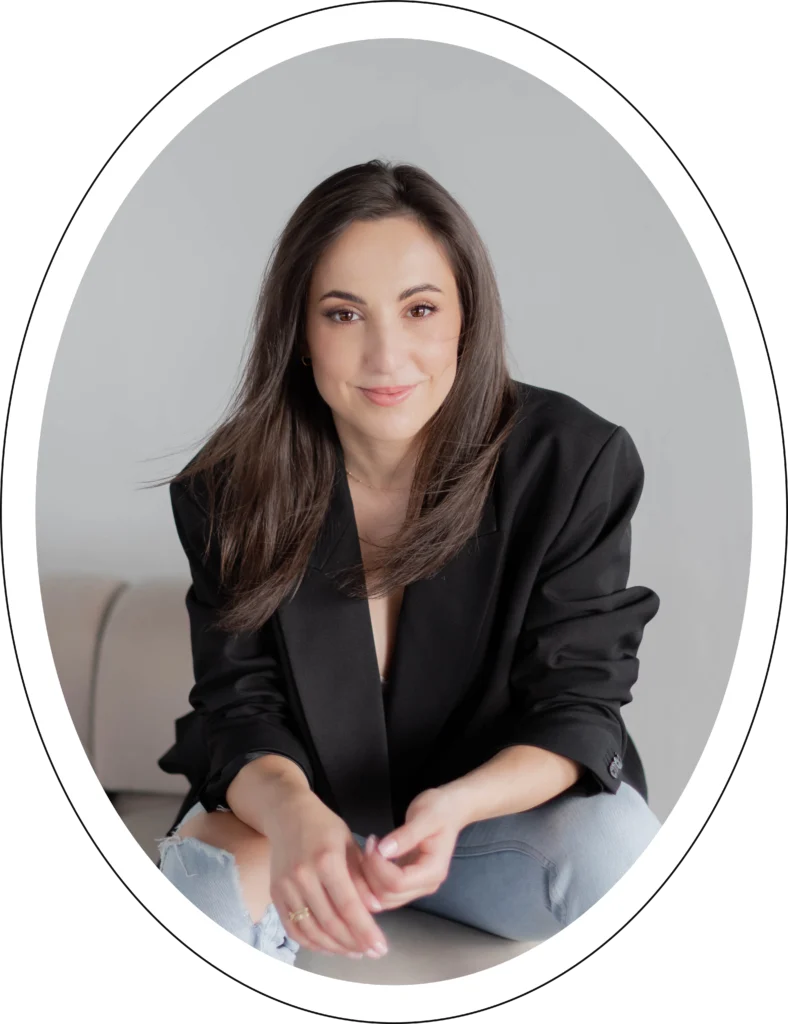 Młoda kobieta w ciemnych włosach siedzi na kremowej kanapie. Ma na sobie czarną marynarkę i jeansy.