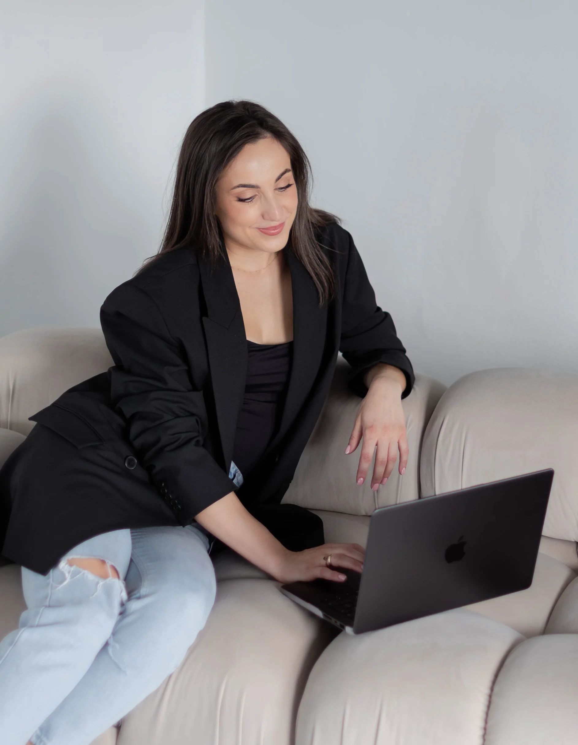 Młoda kobieta w ciemnych włosach siedzi na kremowej kanapie i przegląda coś na laptopie. Ma na sobie czarną marynarkę i jeansy.