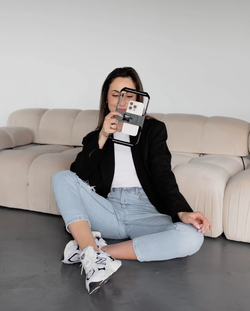 Młoda kobieta w ciemnych włosach stoi i uśmiecha się do kamery. Ma na sobie czarną marynarkę i jeansy. Trzyma w rękach telefon w obudowie do nagrywania reelsów.