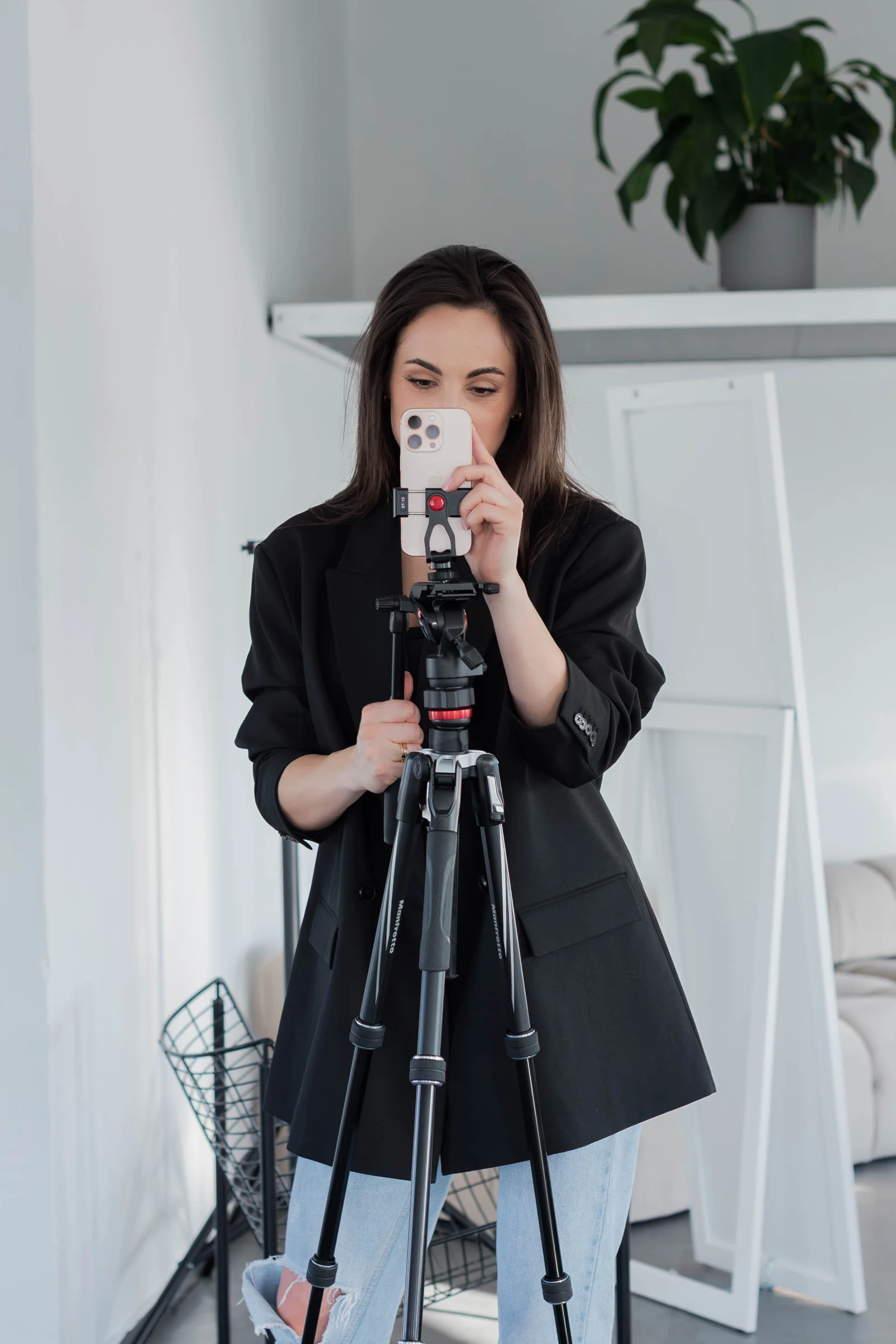 Młoda kobieta w ciemnych włosach stoi i skupia swój wzrok na telefonie przyczepionym do statywu. Ma na sobie czarną marynarkę i jeansy.