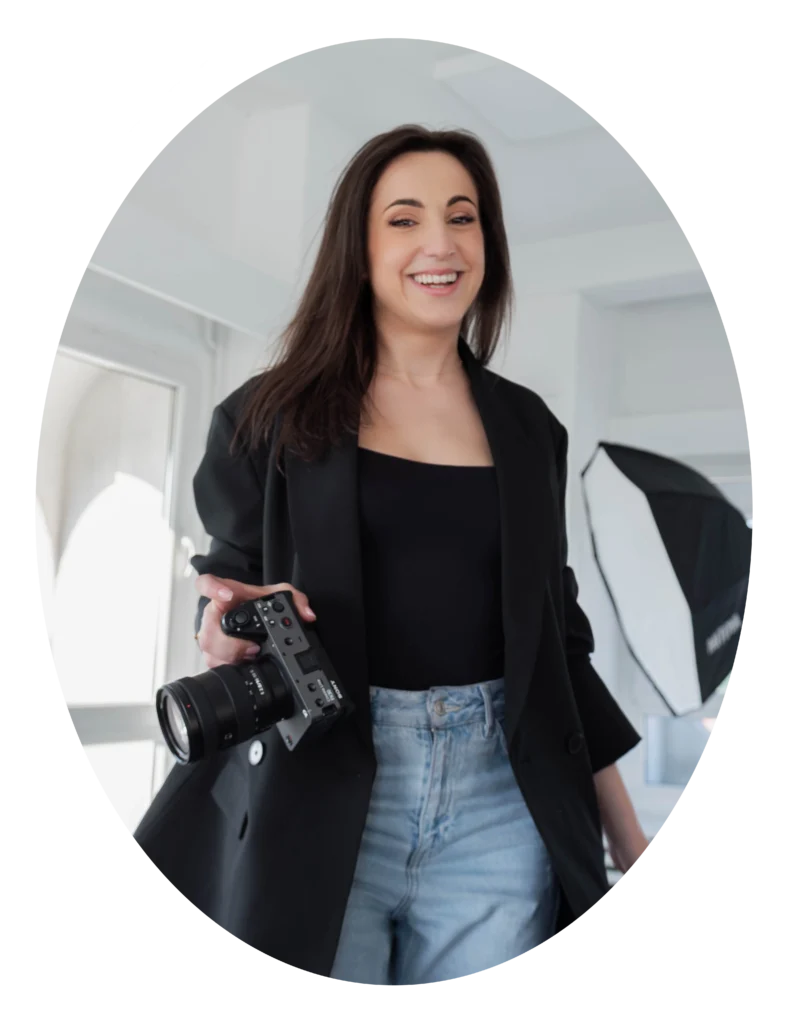 Młoda kobieta w ciemnych włosach stoi i uśmiecha się do kamery. Ma na sobie czarną marynarkę i jeansy. Trzyma w dłoni aparat.